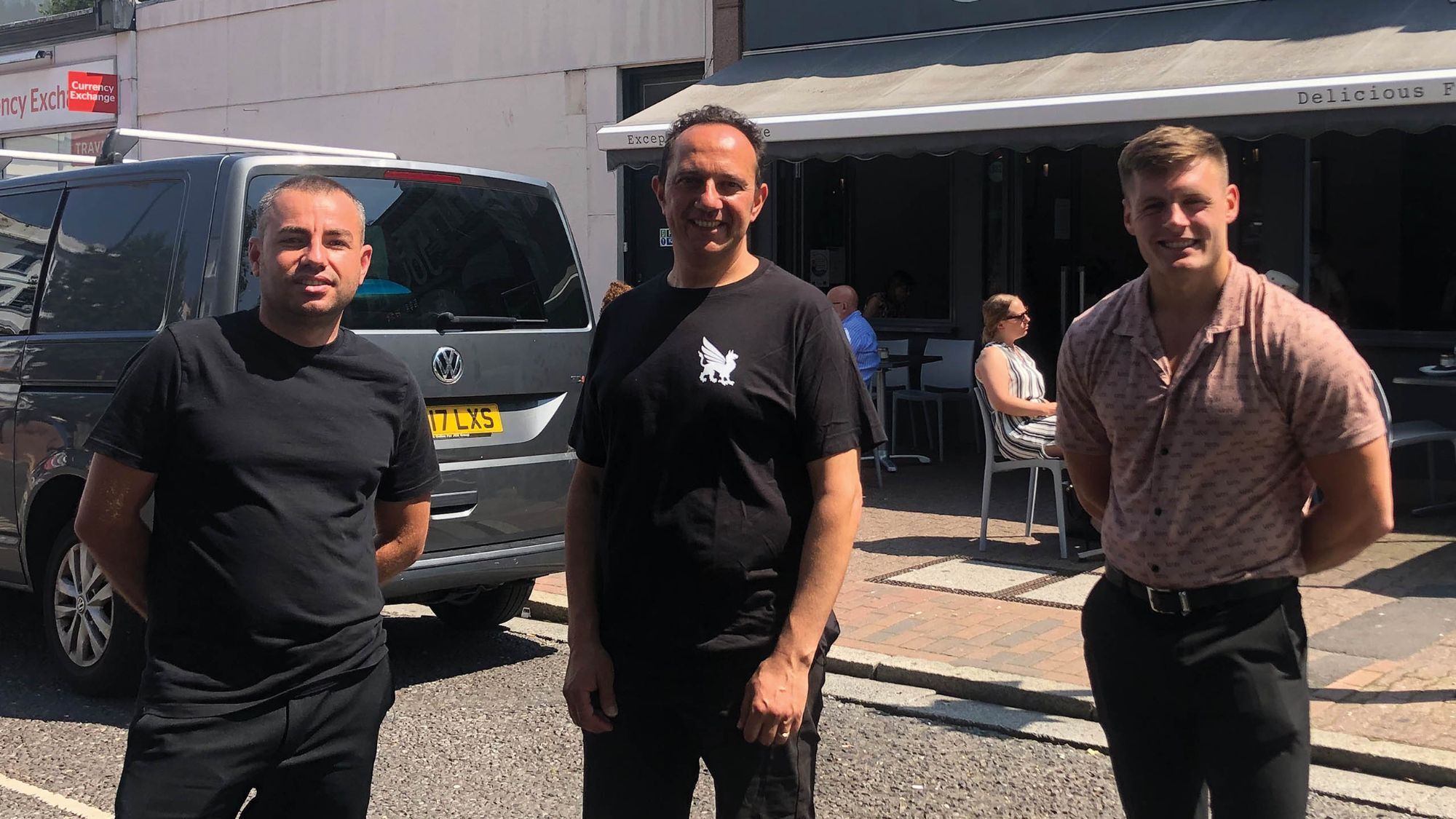 Three men smiling outside a café on a sunny town centre street