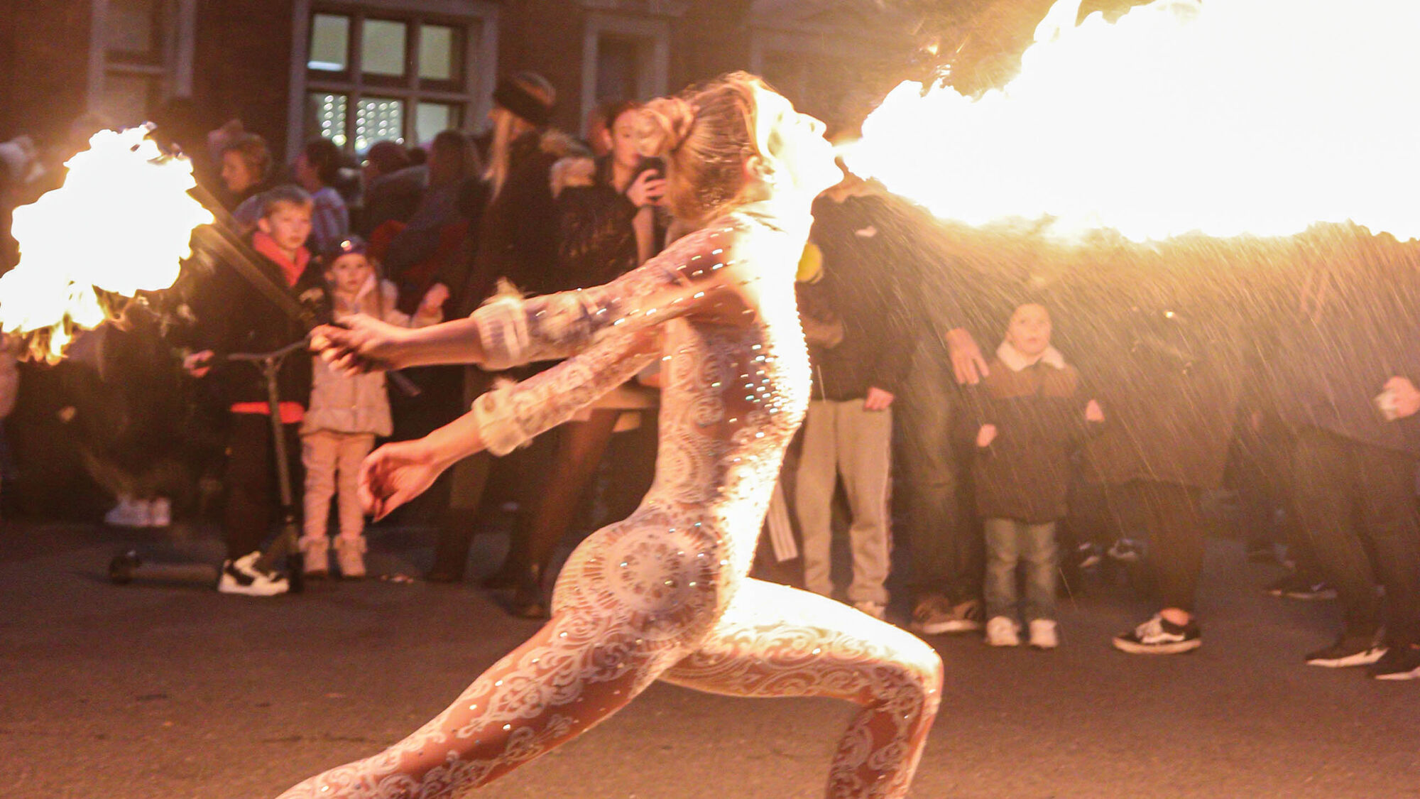 A fire performer in a sparkling white costume breathing fire in front of a crowd on an Eastbourne street at night
