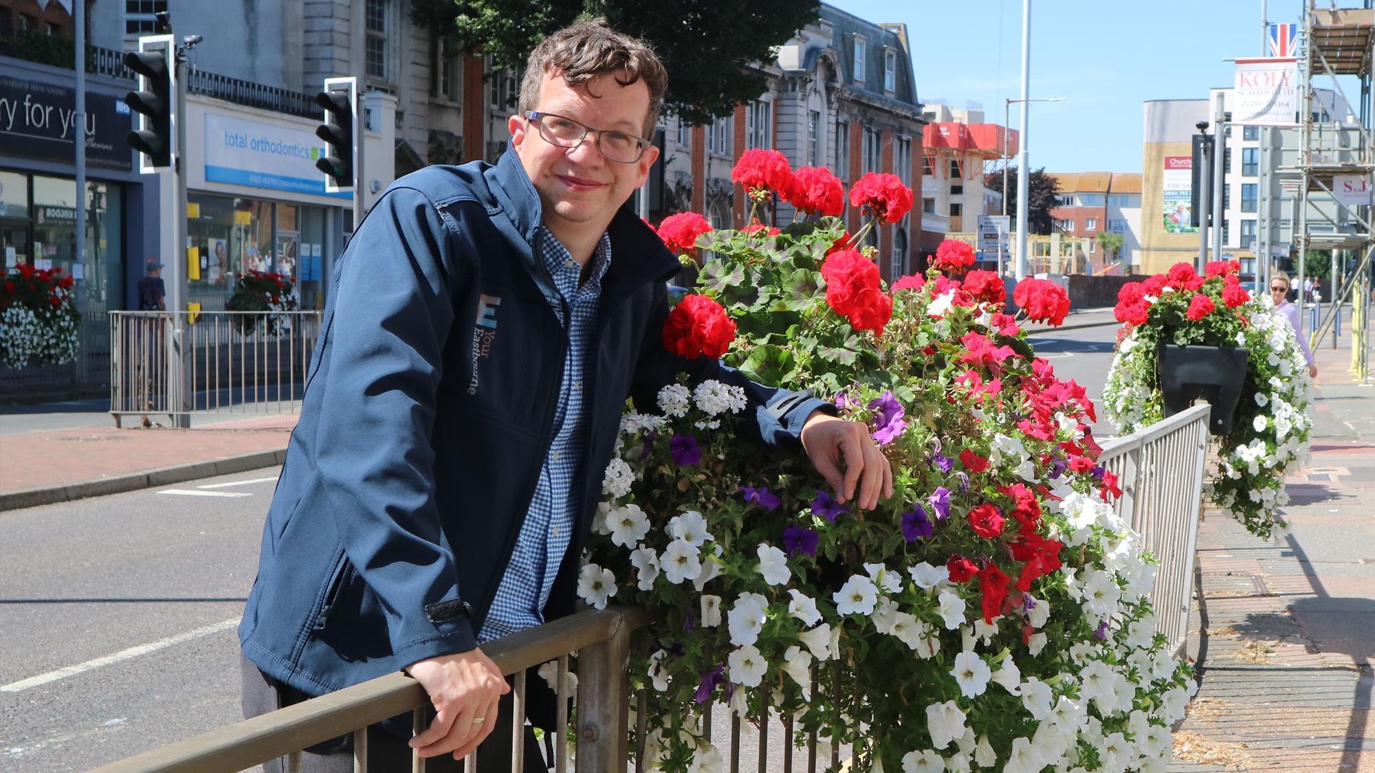 A smiling member of the Your Eastbourne BID team beside a planter overflowing with red, white and purple flowers on a sunny town centre street